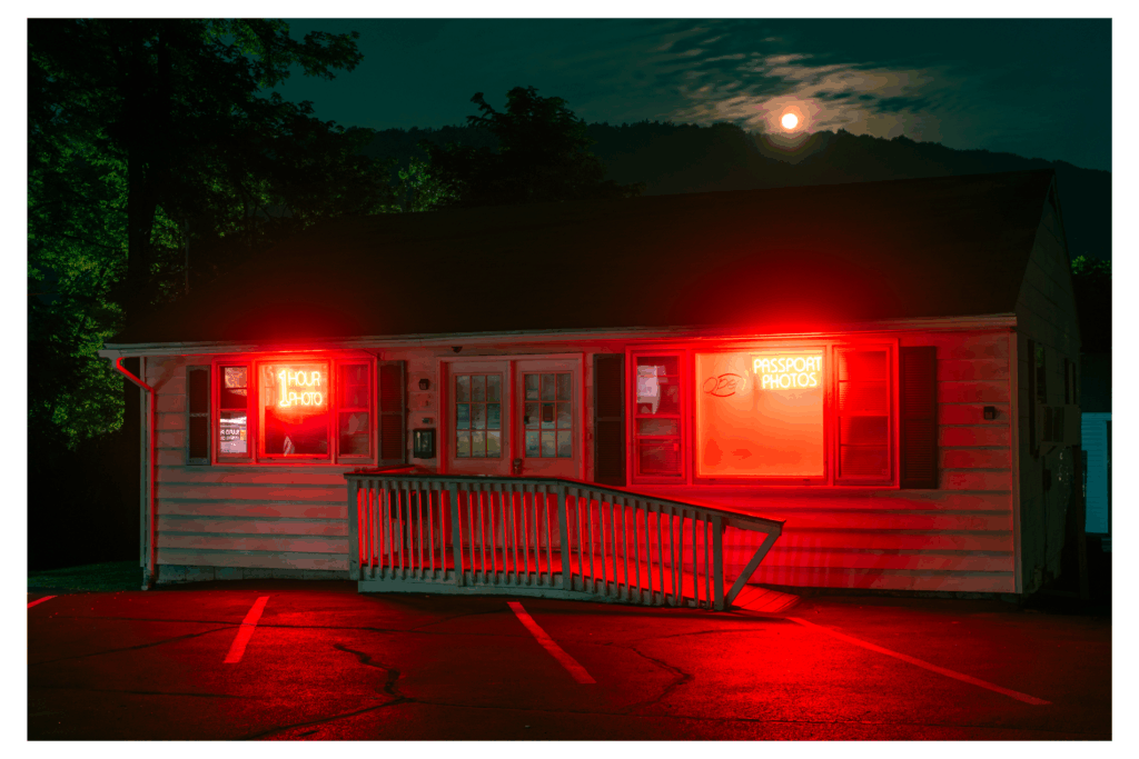 A moody photograph of a small photo developing shop under a rising moon on rain-slicked streets.